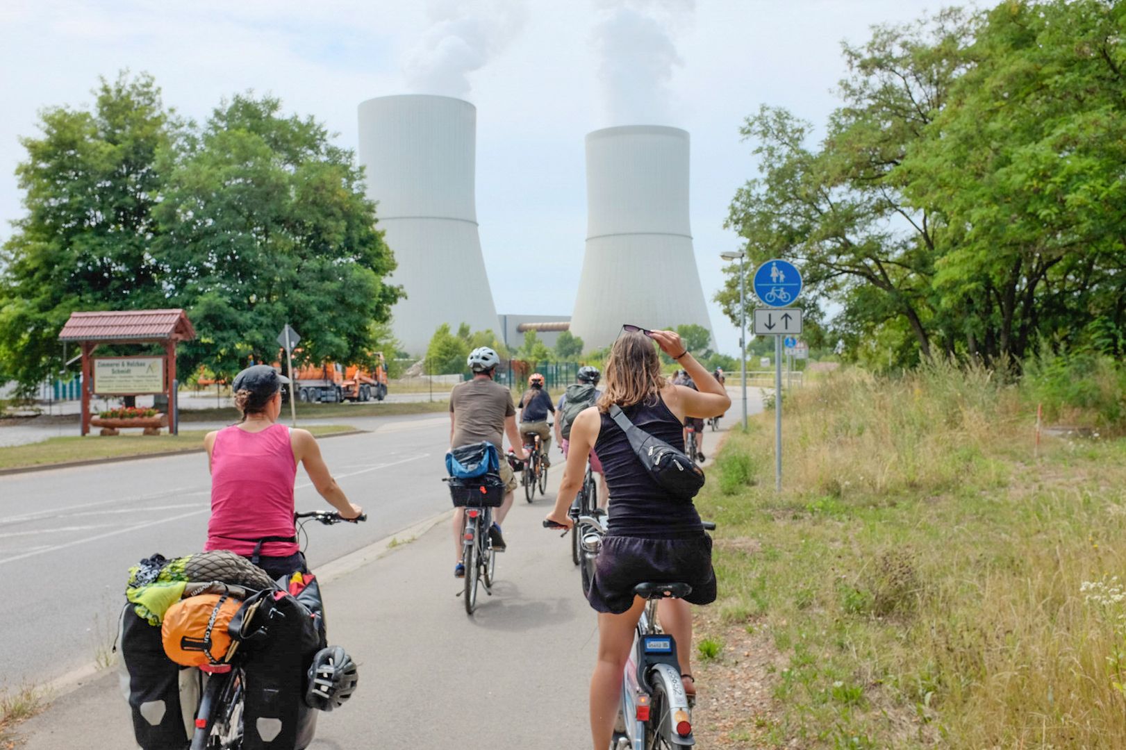Eine Gruppe von Fahrradfaher*innen fahren auf einer Straße. Im Hintergrund sieht man die Türme des Kohlekraftwerks Lippendorf.